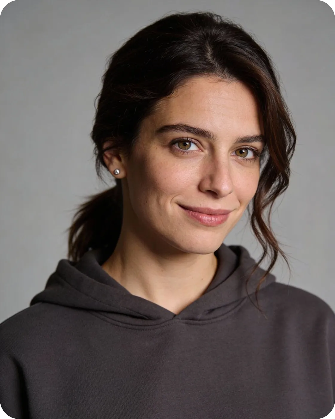 Smiling woman with brown hair tied back wearing a dark hoodie and stud earrings against a neutral background.
