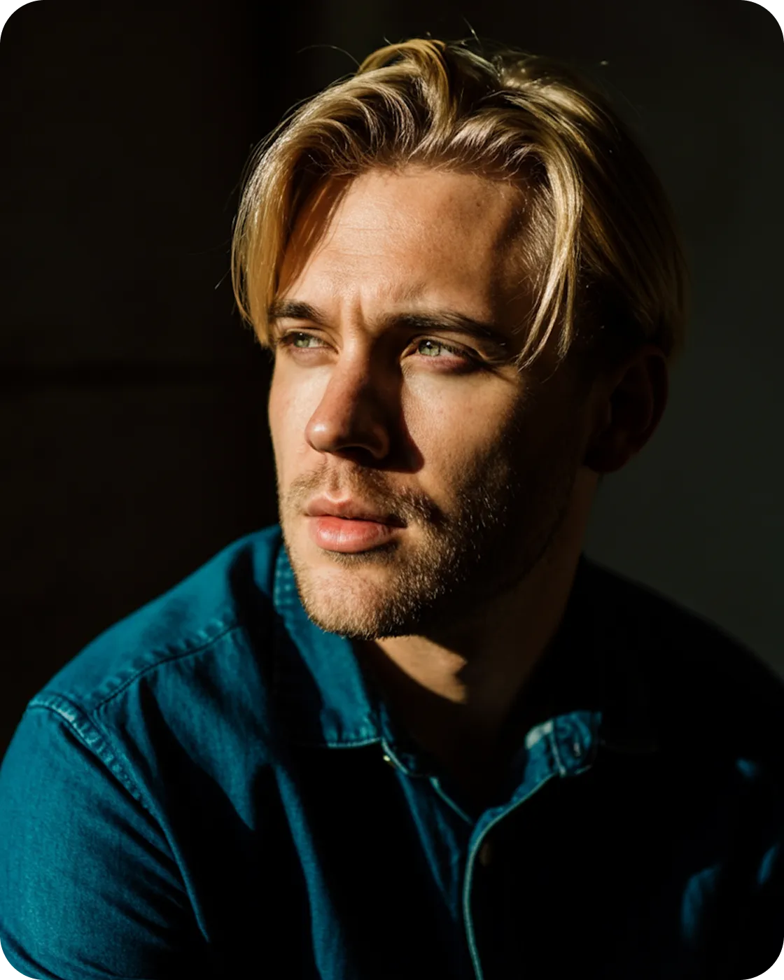 Blond man with light beard wearing a blue shirt, looking thoughtfully to the side in a dark setting with dramatic lighting.