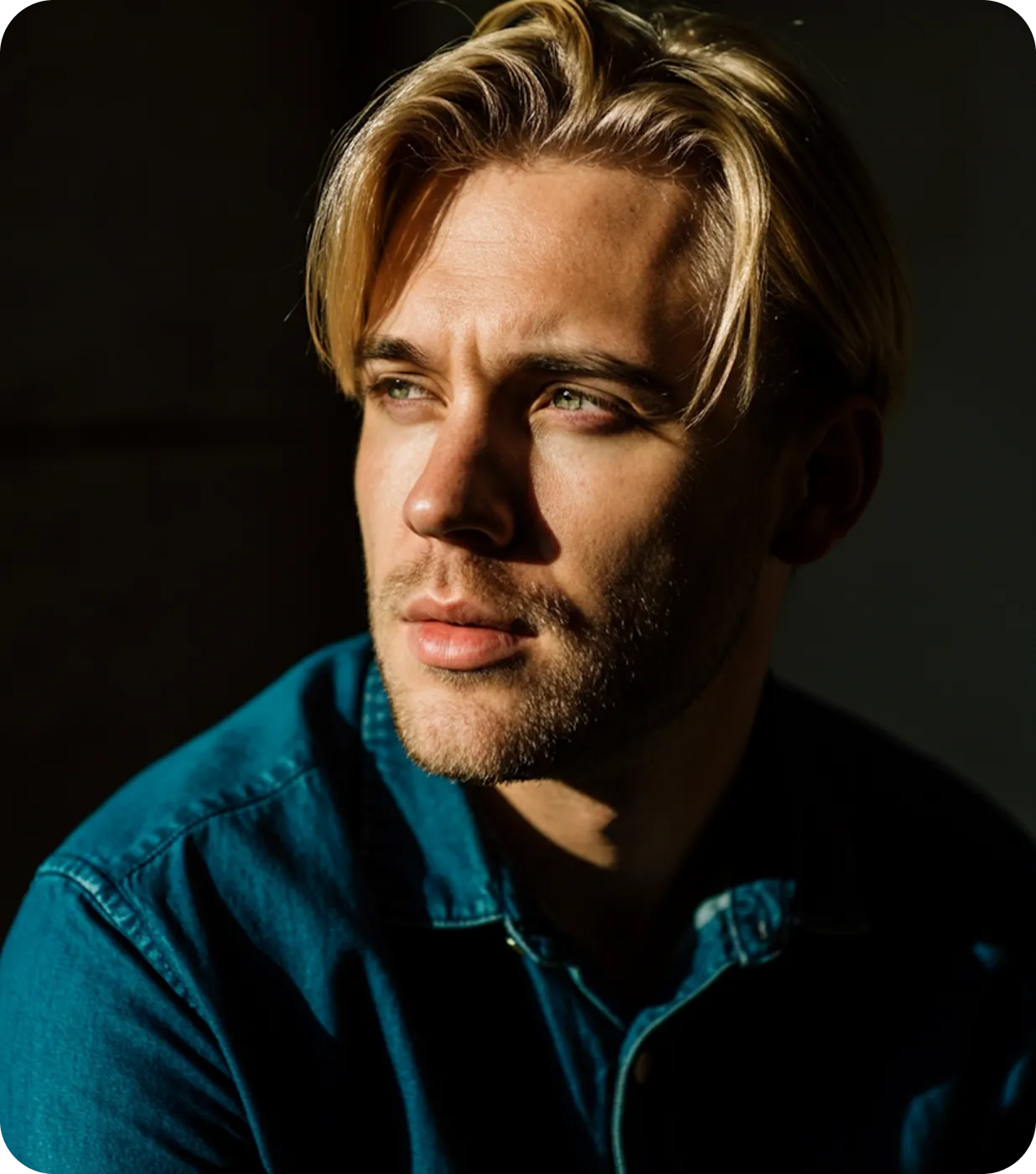 Young man with blonde hair and green eyes wearing a denim shirt, looking thoughtfully to the side with sunlight on his face.