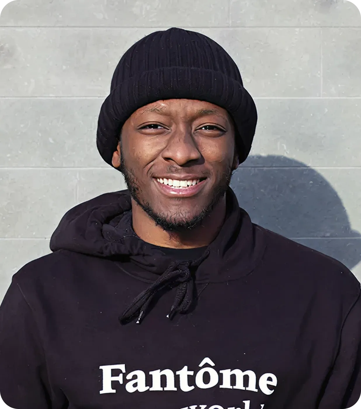 Smiling man wearing a black knit hat and a black hoodie with 'Fantôme' written on it, standing against a light gray wall.